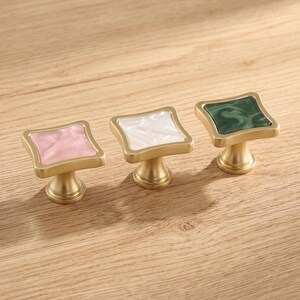 May include: Three square cabinet knobs with a brushed gold finish and a central inlay. The knobs feature a marbled resin surface in light pink, white, and dark green, each set within a square frame with slightly curved edges and a pedestal base.