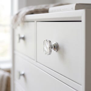 May include: A clover-shaped cabinet knob featuring a polished silver-tone metal frame and a white iridescent mother-of-pearl inlay. The knob is mounted on a white wooden drawer front, with a matching silver-tone knob visible on the drawer below.