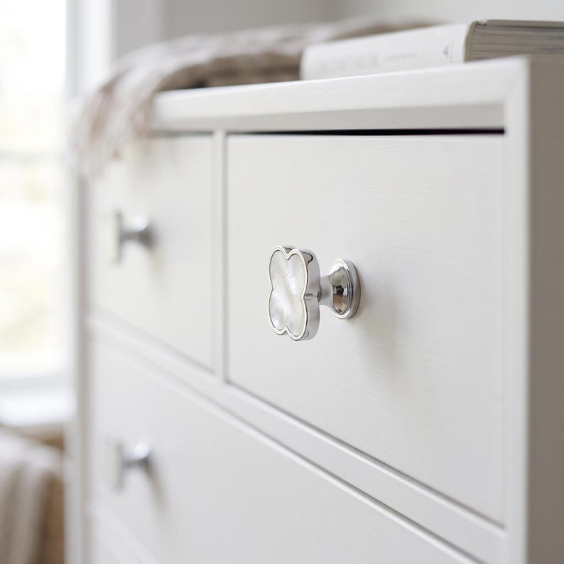 May include: A clover-shaped cabinet knob featuring a polished silver-tone metal frame and a white iridescent mother-of-pearl inlay. The knob is mounted on a white wooden drawer front, with a matching silver-tone knob visible on the drawer below.