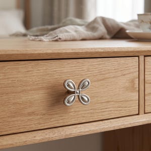 May include: A light-colored wooden drawer featuring a silver-toned metal knob shaped like a four-petaled flower. The knob has a smooth, reflective finish with a small square detail at its center, mounted on the front of the horizontal drawer.