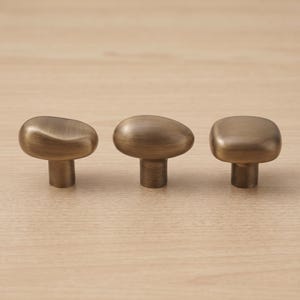 Op de afbeelding: Drie kastknoppen van geborsteld messing, elk met een eigen organische vorm. De knoppen hebben een glad, afgerond bovenoppervlak met een metalen afwerking en een cilindrische basis, gerangschikt in een horizontale rij op een vlak.