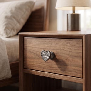 May include: A heart-shaped drawer knob featuring a metallic silver frame filled with numerous small, clear, faceted crystals. The knob is attached to the front of a wooden drawer, showcasing a reflective, textured surface within the heart shape.