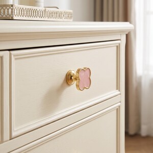 May include: A clover-shaped drawer knob featuring a polished gold-tone metal base and a smooth, light pink center inlay. The knob is mounted on a cream-colored wooden drawer front with a recessed panel design and a visible wood grain texture.