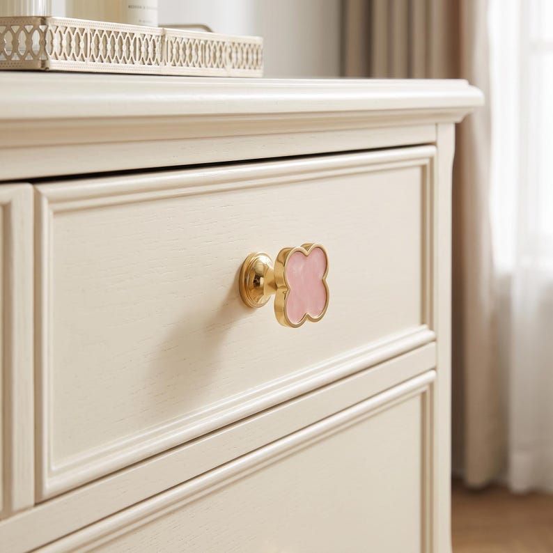 May include: A clover-shaped drawer knob featuring a polished gold-tone metal base and a smooth, light pink center inlay. The knob is mounted on a cream-colored wooden drawer front with a recessed panel design and a visible wood grain texture.