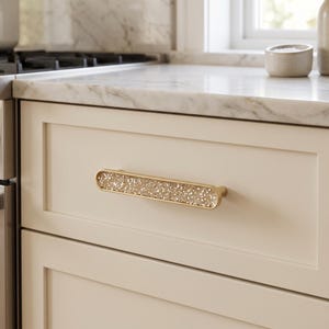 May include: A horizontal drawer pull with a metallic gold finish and a central inlay of clear, textured, crystalline material. The handle is mounted on a cream-colored cabinet drawer featuring a recessed panel design in a kitchen setting.