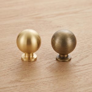 May include: Two spherical cabinet knobs with a circular base. One knob features a smooth, bright brushed gold finish, while the other knob displays a darker, antiqued brass finish with visible horizontal brushed texture lines on the surface.