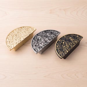 Puede incluir: Tres tiradores de armario semicirculares con un patrón de nubes en relieve. El conjunto incluye un tirador dorado, uno plateado y uno negro con reflejos dorados en las superficies de las nubes, todos con un acabado liso y uniforme.