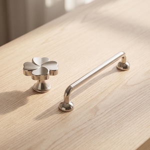 Op de afbeelding: Een zilverkleurige meubelknop in de vorm van een klavertjevier en een bijpassende zilverkleurige meubelgreep met een glad, afgerond stangontwerp en twee ronde bevestigingsvoeten, beide met een gepolijste, reflecterende afwerking.