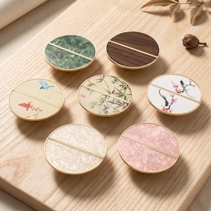 May include: A collection of seven circular cabinet knobs with gold-toned metal rims and a central horizontal bar. The designs include green and pink marbled patterns, wood grain, cream, and white surfaces featuring painted birds and bamboo motifs.