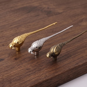 May include: Three bird-shaped cabinet knobs in gold, silver, and antique bronze finishes. Each knob features a detailed bird body with a long, slender tail and a small cylindrical mounting post on the underside for attachment to furniture.