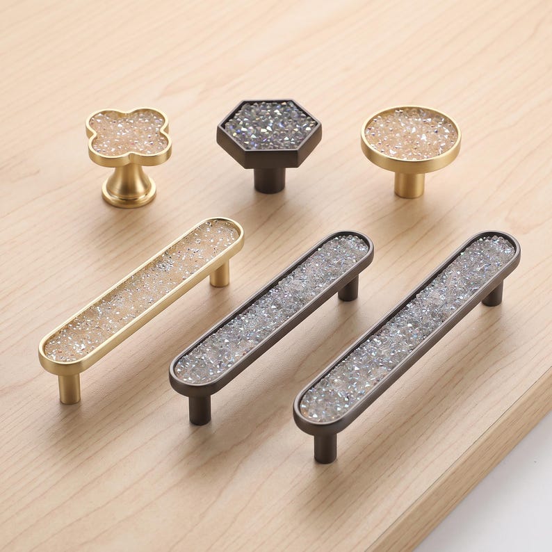 May include: A collection of cabinet hardware including three knobs and three drawer pulls. The knobs feature clover, hexagonal, and circular shapes with gold or dark metal finishes and inlaid crushed crystal textures in clear or champagne tones.