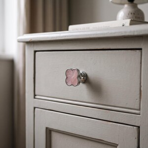 May include: A clover-shaped drawer knob featuring a polished silver-tone metal rim and a smooth, light pink stone inlay. The knob is attached to the front of a light gray wooden drawer, which is part of a small cabinet with a flat top surface.