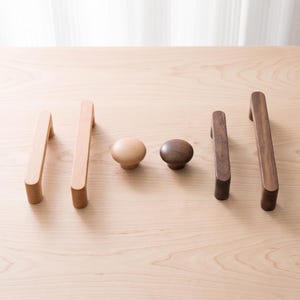 Puede incluir: Una colección de seis piezas de herrajes de madera para muebles que incluye dos tiradores arqueados de tono claro, dos tiradores arqueados de tono oscuro y dos pomos redondos, uno en acabado de madera clara y otro en madera oscura.