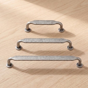 Puede incluir: Tres tiradores de cajón de metal dispuestos en una fila vertical, cada uno con un acabado plateado oscuro desgastado, una barra central plana y rectangular, y extremos redondeados que se curvan hacia bases de montaje circulares con orificios.