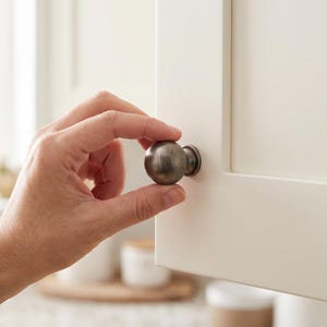 以下が含まれることがあります： 滑らかで丸みを帯びた表面と小さな円形のベースプレートを備えた、つや消しメタリック仕上げの球形キャビネットノブ。シンプルでニュートラルなデザインのオフホワイトのキャビネットドアパネルに取り付けられています。