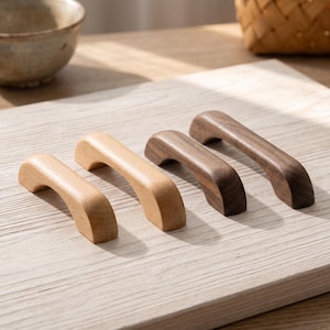 May include: Four wooden cabinet handles arranged in a row. Two handles are made of light-colored wood with a smooth finish, and two handles are made of dark-colored wood with a visible grain. Each handle features a curved, arch-like shape.