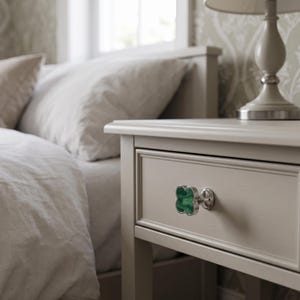 May include: A clover-shaped drawer knob featuring a vibrant green malachite-style inlay with a polished silver-tone metal border and a circular base, attached to a light beige wooden nightstand drawer front.