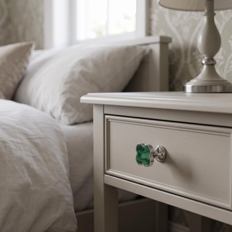 May include: A clover-shaped drawer knob featuring a vibrant green malachite-style inlay with a polished silver-tone metal border and a circular base, attached to a light beige wooden nightstand drawer front.