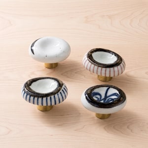 May include: Four round ceramic cabinet knobs with brass-coloured metal bases. One knob is white with a black crescent, one has red vertical stripes, one has blue vertical stripes, and one features a blue painted design, all with dark rims.