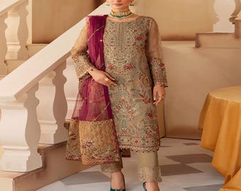 May include: A beige embroidered salwar kameez ensemble with a matching dupatta. The outfit features intricate floral patterns in red, green, and gold. The ensemble includes a long-sleeved tunic, matching pants, and a sheer maroon dupatta.