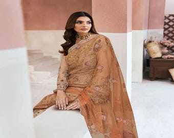 May include: A woman wearing a tan-colored embroidered outfit with a sheer overlay. The outfit includes a long-sleeved top and matching pants. The sheer overlay drapes over the shoulders and arms. The background features a neutral color palette.