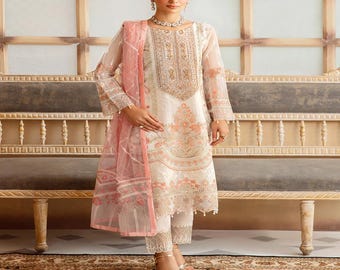 May include: An ivory-colored embroidered outfit with a matching sheer pink scarf. The ensemble includes a long-sleeved tunic, matching pants, and intricate floral embroidery. The outfit is worn with gold sandals and a delicate necklace.