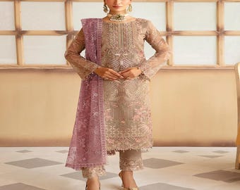 May include: A formal ensemble featuring a long-sleeved, embroidered dress in shades of beige and pink, paired with matching pants. A sheer, mauve scarf drapes over the shoulder. The outfit is accessorized with gold-colored heels and jewelry.