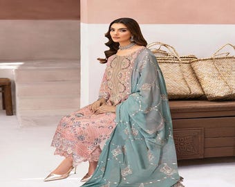 May include: A woman wearing a pink and blue embroidered ensemble. The outfit includes a long-sleeved top and matching pants, paired with a sheer blue scarf. Gold heels complete the look. Woven baskets sit in the background.