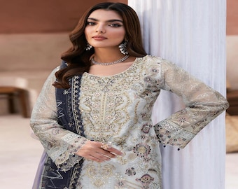 May include: A light-colored, embroidered dress with long sleeves. The dress features intricate floral and geometric patterns in shades of gold, purple, and white. A dark blue scarf is draped over the shoulder. The model is wearing jewelry.