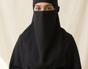 May include: A solid black niqab featuring a layered design that covers the head and face, leaving the eyes visible. The fabric is opaque and drapes over the shoulders, creating a uniform appearance with a smooth texture and clean, straight edges.