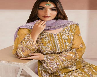 May include: Mustard-colored embroidered tunic and matching pants with a sheer overlay. The tunic features intricate floral and geometric patterns in shades of white and purple. A matching lavender scarf is draped around the neck. A traditional headpiece with green accents is worn.