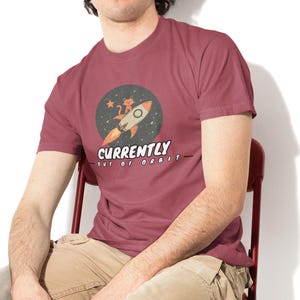 Könnte beinhalten: Burgunderfarbenes T-Shirt mit Rundhalsausschnitt und einem Aufdruck einer Rakete mit einer Katze und Sternen in einem Kreis. Der Text "CURRENTLY out of ORBIT" steht darunter. Das T-Shirt wird von einer Person getragen.