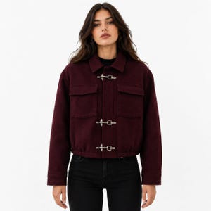 May include: A deep burgundy cropped jacket featuring a classic collar, long sleeves, and two chest pockets with flaps. The front closure consists of three silver-toned metal toggle clasps. The hem is finished with an elasticized band for a snug fit.