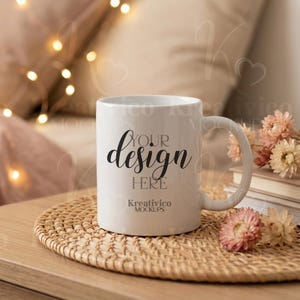 May include: A white ceramic mug with the text "YOUR design HERE" in black script. The mug sits on a woven coaster, with dried flowers and books in the background. The mug has a curved handle and the words "Kreativico MOCKUPS" at the bottom.