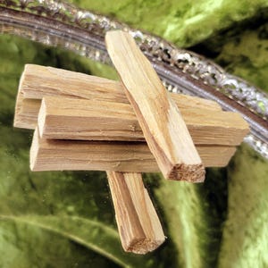 May include: A collection of light brown Palo Santo sticks, arranged on a reflective surface. The sticks have a natural wood grain pattern and are stacked in a visually appealing manner. The background features a green, textured fabric and a decorative silver mirror.