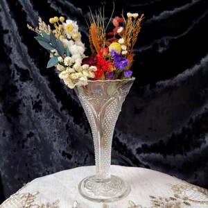 May include: A clear glass vase with a flared top and a textured, diamond-patterned body. The vase holds a colorful arrangement of dried flowers, including white, yellow, red, and purple blooms, and various dried grasses. The vase sits on a white surface.