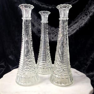 May include: Three clear glass bud vases with a tapered, conical shape. Each vase features a textured, pressed glass pattern with geometric diamond and starburst motifs, a flared rim, and a smooth, circular base for stability and display.