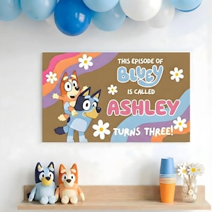 Puede incluir: Un letrero de lona marrón con el texto "THIS EPISODE OF BLUEY IS CALLED ASHLEY TURNS THREE!" presenta personajes de dibujos animados y detalles de margaritas. Globos azules y naranjas en el fondo, con peluches y decoraciones en un estante debajo.