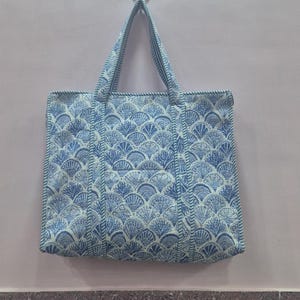 Könnte beinhalten: Eine blau-weiße Tragetasche mit einem sich wiederholenden fächerförmigen Muster. Die Tasche hat zwei Griffe und hängt an einer hellrosa Wand. Die Tasche ist aus einem strukturierten Stoff gefertigt.