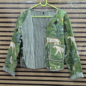 Puede incluir: Una chaqueta acolchada con base verde y estampado de selva con leopardos, tucanes y palmeras. La chaqueta tiene cuello en V, mangas largas con puños a rayas, un bolsillo delantero de parche y un forro interior a rayas blancas y negras.