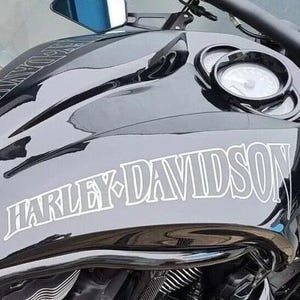 Könnte beinhalten: Nahaufnahme eines schwarzen Harley-Davidson Motorradtanks. Der Tank hat eine glänzende Oberfläche mit dem Harley-Davidson-Logo in Weiß. Die Anzeigen und der Lenker des Motorrads sind sichtbar.