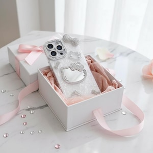 May include: A clear phone case with a sparkling Hello Kitty design, presented in a white gift box with a pink ribbon. The case features a bow and other embellishments made of rhinestones. The box is lined with pink tissue paper.