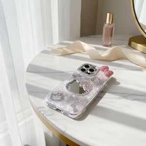 May include: A clear phone case with a pearlescent design, adorned with sparkling rhinestones and a pink bow. The case features a mirror and several Hello Kitty-themed embellishments. The case is on a white marble surface.