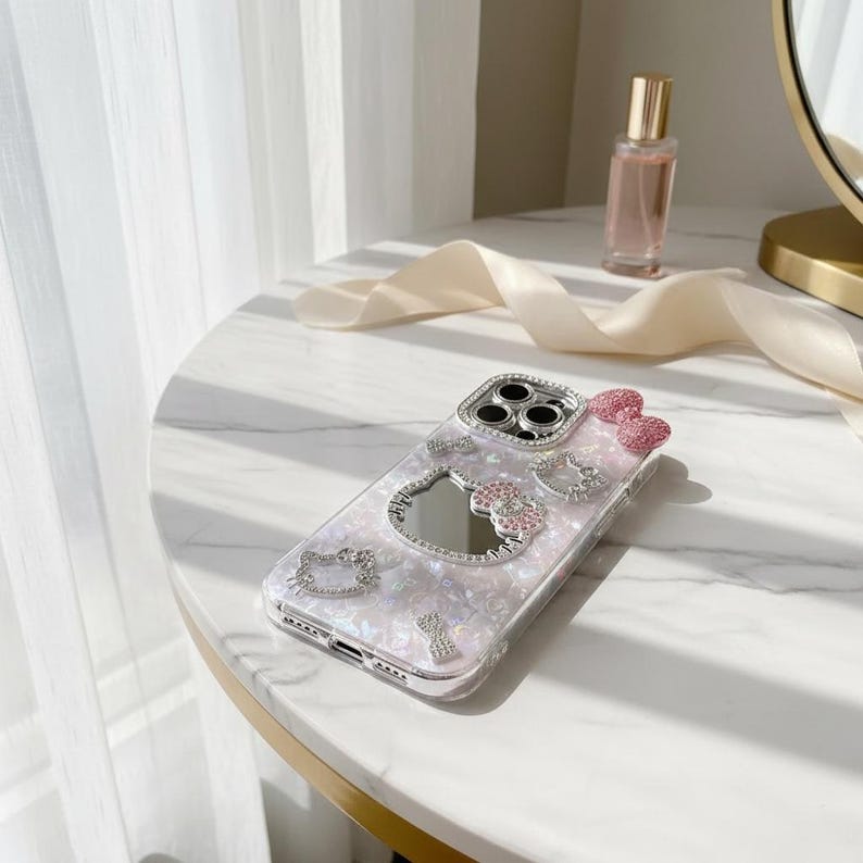 May include: A clear phone case with a pearlescent design, adorned with sparkling rhinestones and a pink bow. The case features a mirror and several Hello Kitty-themed embellishments. The case is on a white marble surface.