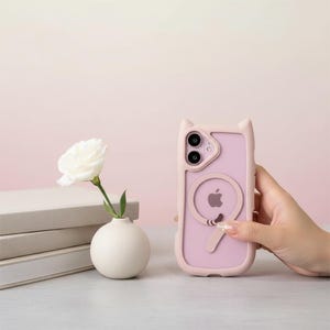 May include: A pink phone case with cat ears and a circular design featuring the Apple logo. The case is held in front of a soft pink background. A white rose in a small white vase is in the background.
