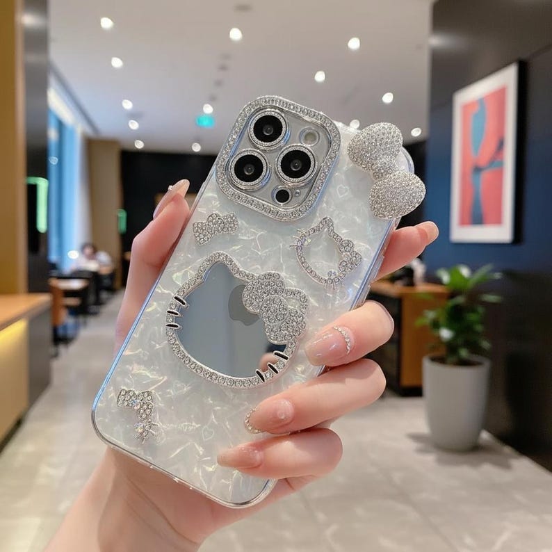 May include: A white and silver phone case with a pearlized finish, featuring a Hello Kitty design and bow accents, embellished with sparkling rhinestones. The case has a camera lens protector with rhinestones.