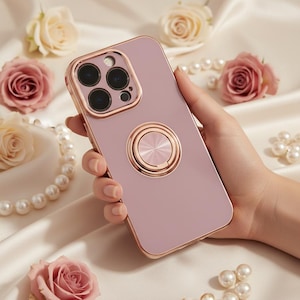 May include: A mauve-colored phone case with a gold-colored ring holder and camera frame. The case is surrounded by roses and pearls. The ring holder is circular and has a textured design. The case is designed for a smartphone.