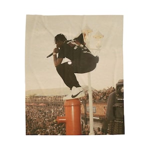 May include: A concert image featuring a performer in a black t-shirt and pants, wearing white sneakers, mid-air on a red fire hydrant. The background shows a large crowd and stage lights.