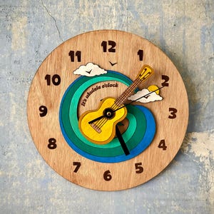May include: Wooden wall clock featuring a ukulele design. The clock face is light brown with brown numerals. A yellow ukulele is centered on a blue, green, and yellow swirl. The text "It's ukulele o'clock" is on the clock face.