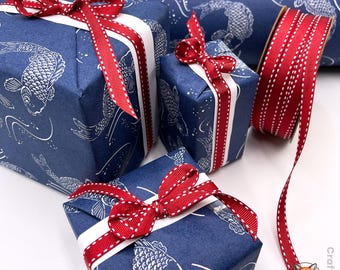 May include: Three gift boxes wrapped in dark blue paper featuring a white koi fish pattern, each tied with a white ribbon and a red ribbon with white stitched edges. A matching roll of red stitched ribbon sits beside the wrapped boxes.